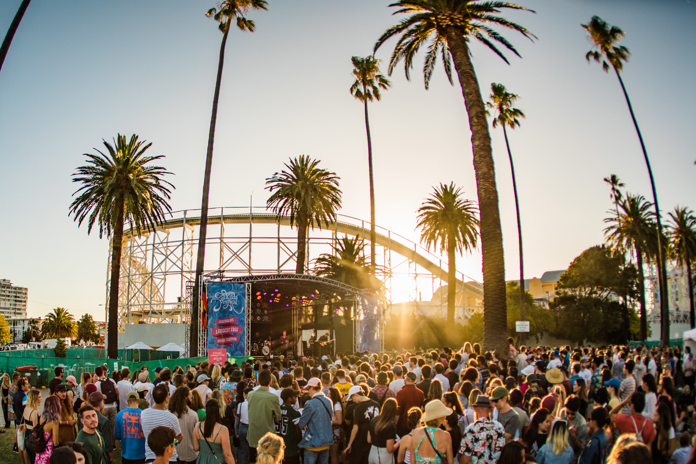A mass of people standing in front of a stage surrounded by palm trees with a rollercoaster in the background
